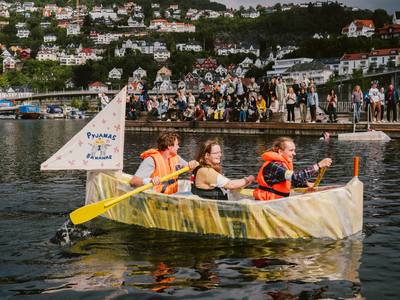 Tre studenter i en hjemmelaget båt på fjorden. Publikum på brygge i bakgrunnen.