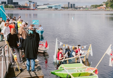 En gjeng med studenter går sammen for å delta i et båt race på KMD. Noen er i vannet på båten, mens flere står på bryggen og ser på. 