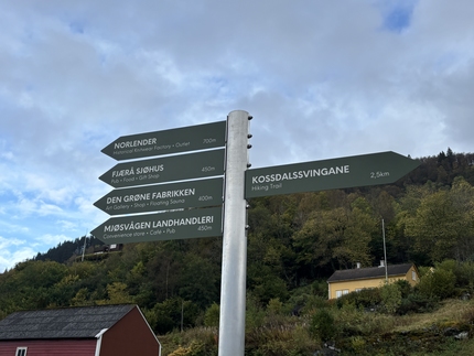 Green signpost with one arrow to the Kossdalssvingane hiking trail, 2.5 km, and four arrows the opposite way to a pub, café, shop and art gallery. Photo