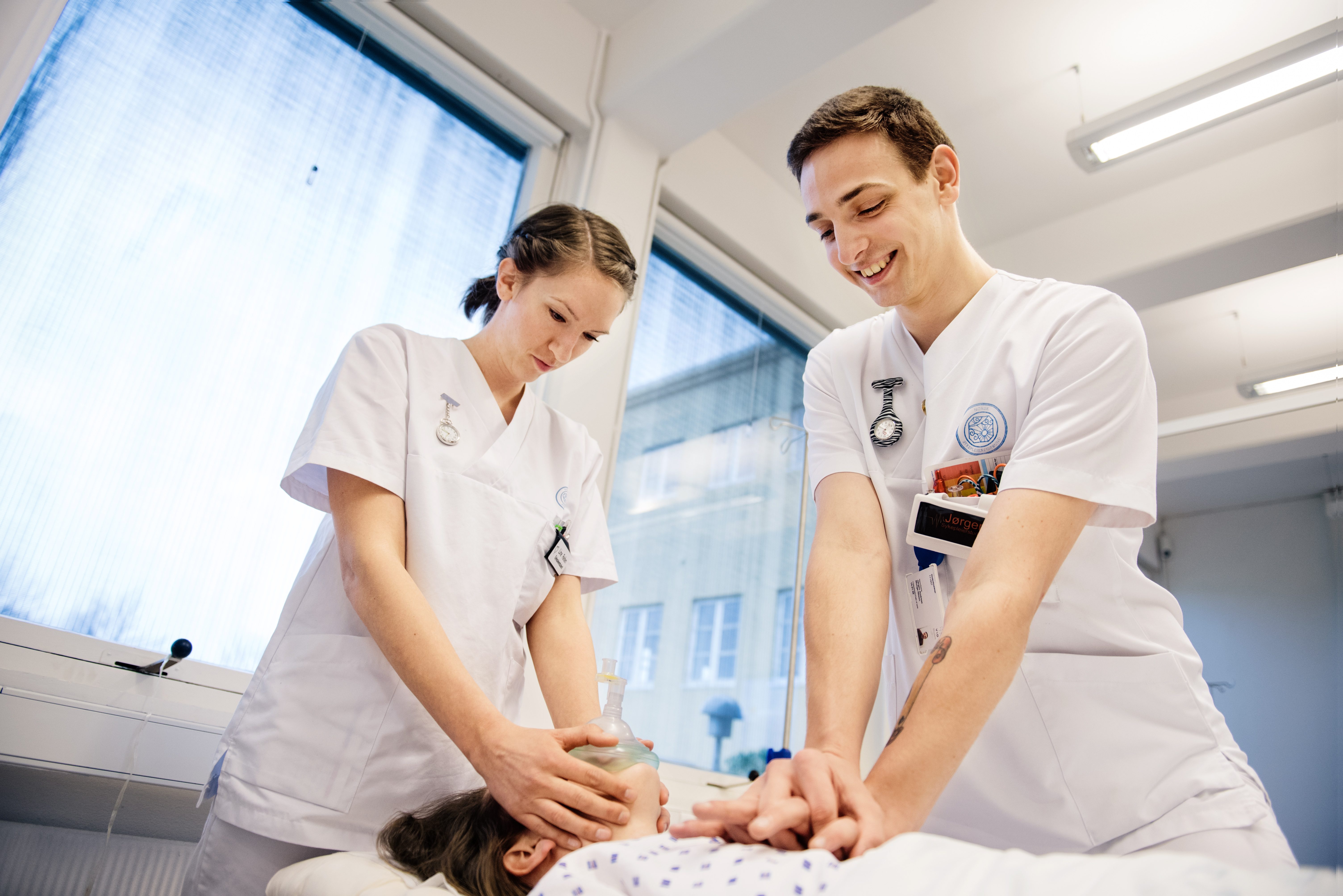 To helsestudenter i hvit uniform jobber med liggende pasient.