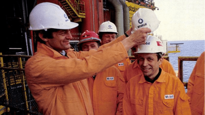 Fire mennesker på en plattform i Nordsjøen i oransje arbeidsdresser. Kollega Kjølv Egeland setter en symbolsk Statoil-hjelm på hodet hans.
