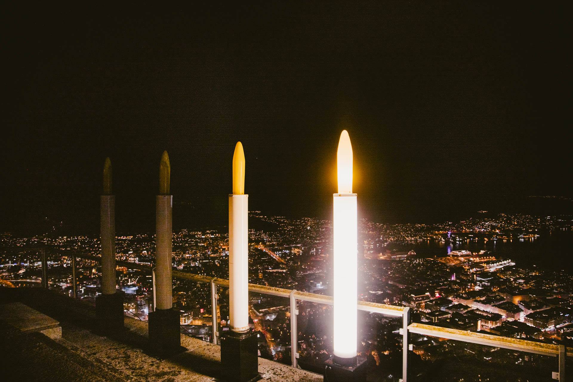 Bildet viser en utendørs adventsstake med fire enorme lys, ett av lysene er tente. I bakgrunnen ses Bergen by.