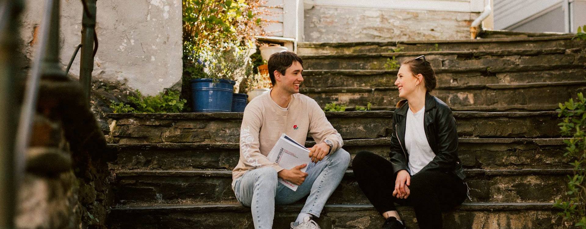 To studenter sitter i en trapp i Bergen og ser på hverandre og smiler.