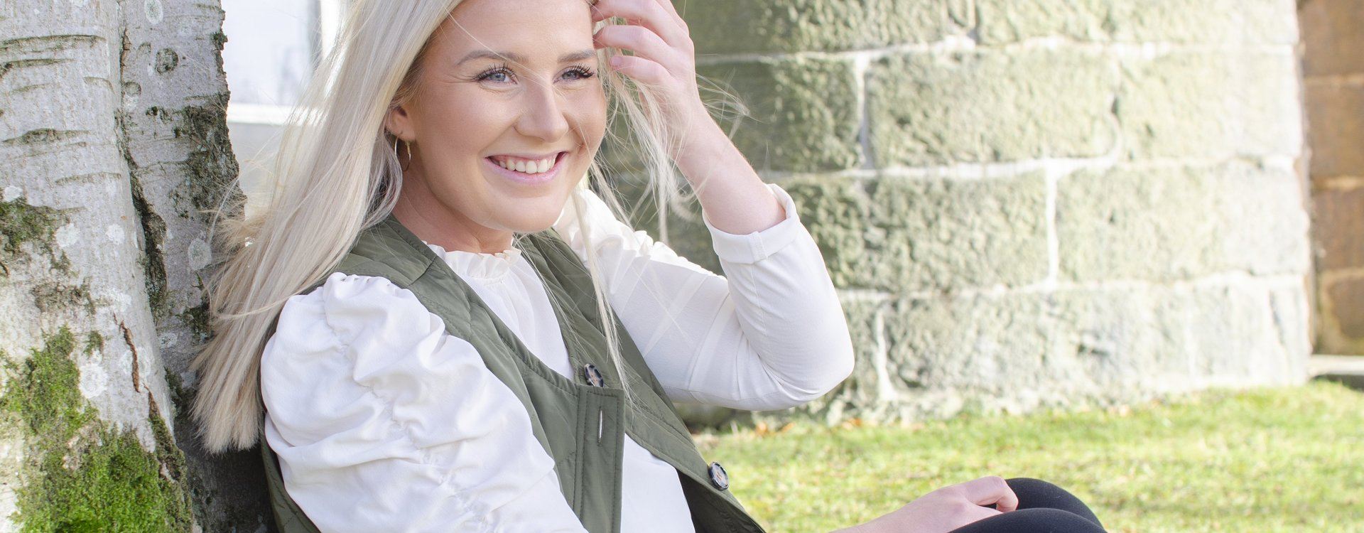 Susan sitter utendørs inntil en trestamme. Nærbilde. Smiler.