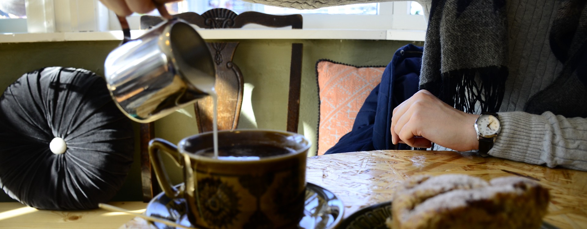 A table with a coffee cup and a plate with cake, a person is pouring cream into coffee.