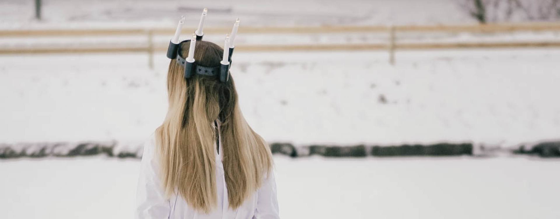 Girl with long blond hair wearing a crown of candles on her head. Seen from behind.