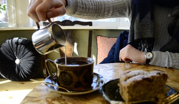 A table with a coffee cup and a plate with cake, a person is pouring cream into coffee.