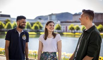Tre studenter smiler og ler, foran Lille Lungegårdsvann i Bergen i strålende solskinn, med fjell i bakgrunnen.