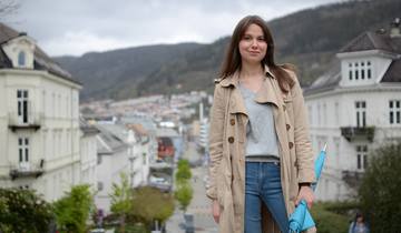 Student i Bergen i halvprofil, med lang kåpe. Paraply i hånden. Utsikt.