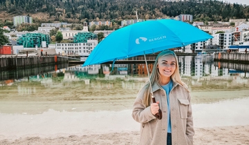 Et bilde: En jente står med en paraply med en sjø i bakgrunnen og smiler